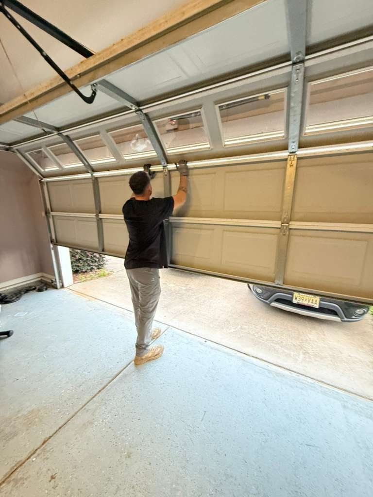 Person standing under a tan sectional garage door raised partway, holding a section by hand, with a car in the driveway beyond