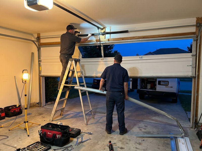 Two technicians in a garage at dusk, one on a stepladder at the ceiling track and one holding a long metal track piece, with a work light and open tool bags on the floor