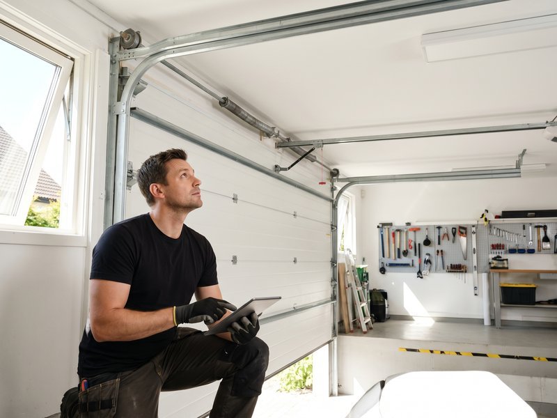 Technician kneeling in a garage and looking up at the ceiling track while holding a tablet