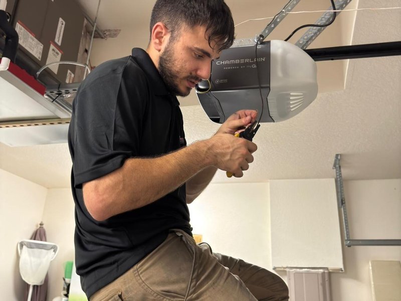 Technician in a black shirt holding a small hand tool beside a Chamberlain opener head mounted on the garage ceiling