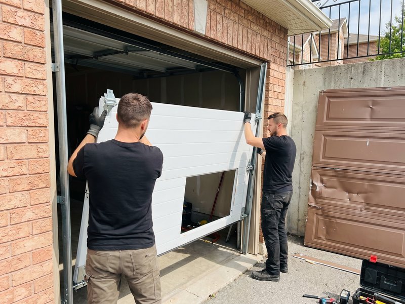 Two technicians lifting a white replacement panel into a brick-front garage door opening